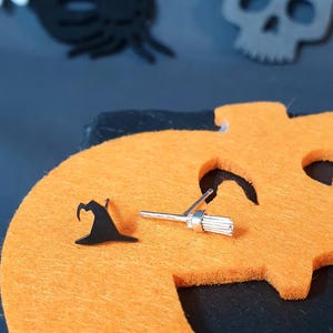 May include: A pair of silver Halloween-themed earrings. One earring features a witch's hat and the other earring features a broom.