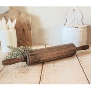 Può includere: Un mattarello vintage in legno con manici marrone scuro e un corpo cilindrico. Il mattarello è esposto su una superficie di legno di colore chiaro, suggerendo un'estetica rustica o da fattoria. Altri oggetti da cucina sono sullo sfondo.