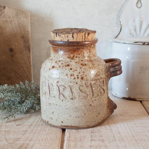 May include: A ceramic jar with a cork stopper. The jar is beige with brown speckles and a brown rim. The word "CERISES" is carved into the front. It has a handle on the side. The jar is on a wooden surface.