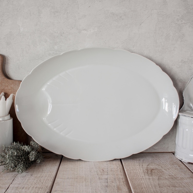 May include: A large, white, oval serving platter with a scalloped edge. The platter has a subtle leaf-like design in the center. The background is a light gray wall and a wooden surface.