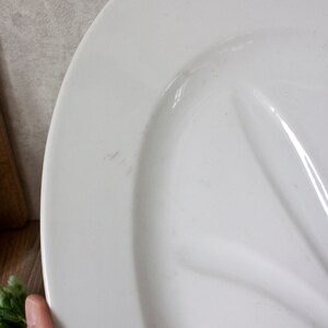 May include: A white oval serving platter with a raised, curved design. The platter is made of ceramic or porcelain and has a glossy finish. The edge of the platter is slightly raised. The background includes a wooden cutting board and greenery.