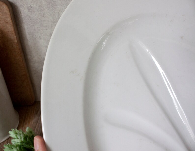 May include: A white oval serving platter with a raised, curved design. The platter is made of ceramic or porcelain and has a glossy finish. The edge of the platter is slightly raised. The background includes a wooden cutting board and greenery.