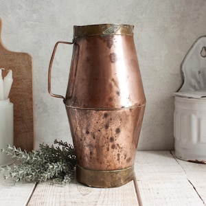 Könnte beinhalten: Ein antiker Kupferkrug mit Messingrand und -fuß. Der Krug hat einen Henkel und eine leicht konische Form. Das Kupfer hat eine reiche, gealterte Patina und steht auf einer weißen Holzoberfläche.