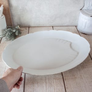 May include: A large, white, oval serving platter with a scalloped edge. The platter features a subtle leaf design on one side. The dish is held by a person's hand, showcasing its size and shape. Ideal for serving food.