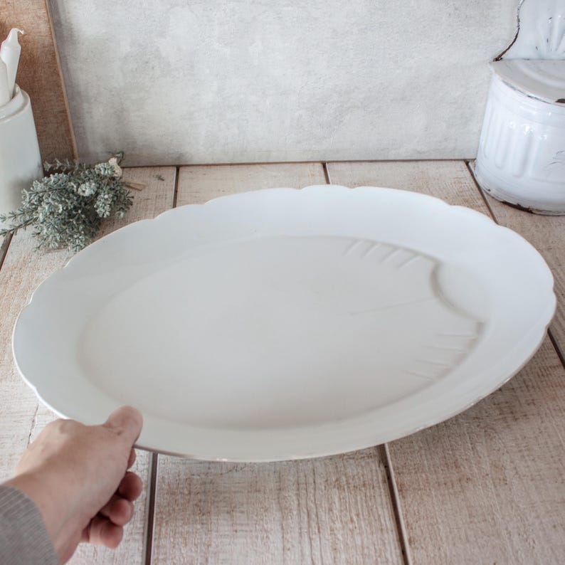 May include: A large, white, oval serving platter with a scalloped edge. The platter features a subtle leaf design on one side. The dish is held by a person's hand, showcasing its size and shape. Ideal for serving food.