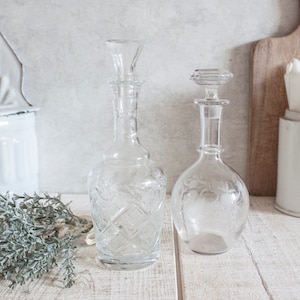May include: Two clear glass decanters with etched designs. The decanters have a round body and a tall, slender neck. The decanters are sitting on a white wooden surface.