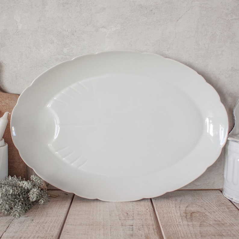 May include: A large, white, oval serving platter with a scalloped edge. The platter has a slightly raised center and a subtle embossed design. The background features a wooden surface and a neutral-toned wall.