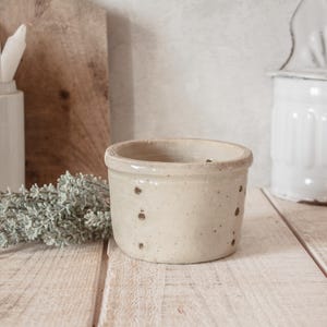 May include: A small, off-white ceramic crock with several small holes around the sides. The crock has a slightly flared rim and sits on a light-colored wooden surface. The background includes a wooden cutting board and a white container.