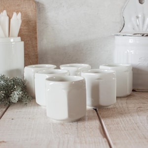Può includere: Una collezione di sei tazze in ceramica bianca con un design geometrico. Le tazze sono disposte su una superficie di legno chiaro, con altri contenitori in ceramica bianca sullo sfondo. Le tazze sono alte circa 7,6 cm.