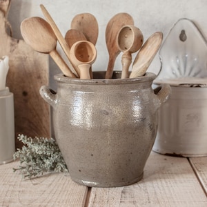 May include: A gray ceramic crock with two side handles holds a collection of various wooden kitchen utensils, including several spoons and two small round scoops with perforated bowls, creating a functional storage display.