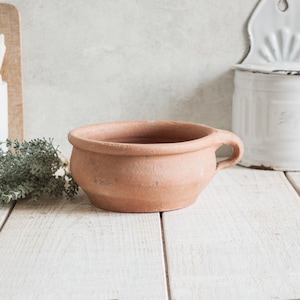 Op de afbeelding: Een terracotta pot met een enkele handgreep. De pot is ongeglazuurd en heeft een rustieke, aardse uitstraling.