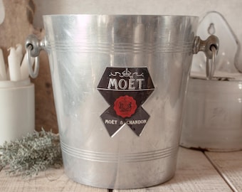 Könnte beinhalten: Ein silberfarbener Champagnerkühler aus Metall mit zwei seitlichen Ringgriffen und einem zentralen schwarzen, schildförmigen Emblem mit dem weißen Schriftzug MOËT und MOËT & CHANDON sowie einem roten runden Siegel mit Krone.