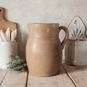 Op de afbeelding: Een rustieke, beige keramische kan met een gebogen handvat. De kan heeft een brede basis en een licht uitlopende rand. De achtergrond omvat een houten snijplank, witte kaarsen in een keramische houder en een decoratieve wandcontainer.