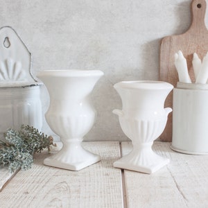 Puede incluir: Dos urnas de cerámica blanca con diseños acanalados y asas. Las urnas están sobre una superficie de madera blanca.
