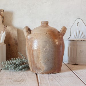 May include: A round, tan-colored stoneware jug with two side handles and a narrow neck. The surface features a mottled, earthy glaze with variations in brown and beige tones, showing a smooth texture with subtle imperfections across the body.