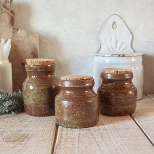 May include: Three small, rustic brown ceramic jars with cork lids. The jars have a speckled, textured finish and a rounded shape. They are arranged on a light-colored wooden surface, with a white enamel container in the background.