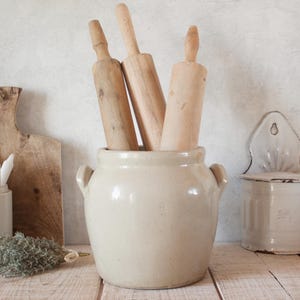 Op de afbeelding: Drie houten deegrollers staan in een crèmekleurige keramische pot met handvatten. De deegrollers hebben verschillende tinten lichtbruin. Een houten snijplank en een witte keramische zoutpot zijn ook zichtbaar.
