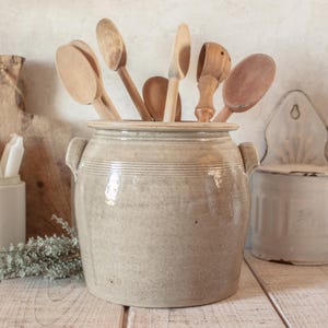 Pode incluir: Um pote de grés grande e cilíndrico em bege claro com duas alças laterais, preenchido com uma variedade de colheres e utensílios de cozinha de madeira. O recipiente de cerâmica apresenta detalhes horizontais estriados no topo.