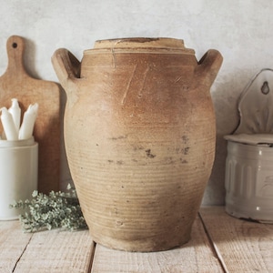 Peut inclure: Un grand pot en céramique de couleur beige antique avec un couvercle et deux poignées. Le pot a une surface texturée et un aspect rustique. D'autres éléments en arrière-plan incluent une planche à découper en bois, des bougies et un petit récipient.