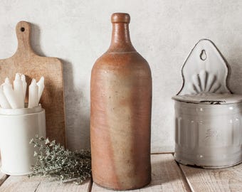 May include: A rustic still life featuring a tall, brown bottle with a narrow neck, a wooden cutting board, a white ceramic container with candles, and a white and gray container. The objects are arranged on a wooden surface.