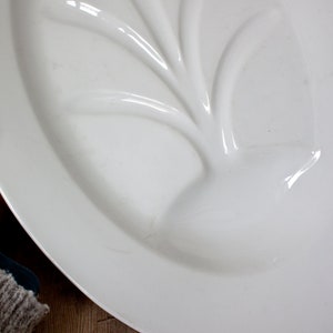 May include: A white, oval-shaped serving platter with a raised, leaf-like design in the center. The platter has a glossy finish and appears to be made of ceramic or porcelain. The design is a subtle, elegant detail.