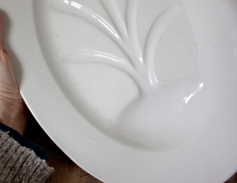 May include: A white, oval-shaped serving platter with a raised, leaf-like design in the center. The platter has a glossy finish and appears to be made of ceramic or porcelain. The design is a subtle, elegant detail.