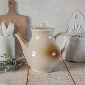 May include: A beige ceramic teapot with a rounded body, spout, and handle. The teapot has a lid and a brown stain on the side. The background includes a wooden cutting board, a white container, and a white wall-mounted container.