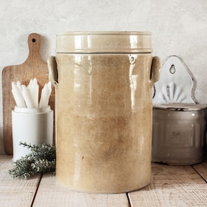 Peut inclure: Un grand pot en céramique beige avec un couvercle et deux poignées latérales. Le pot est cylindrique et a un aspect rustique et vieilli. Une planche à découper en bois, un récipient en céramique blanche avec des bougies blanches et un petit récipient en métal sont en arrière-plan.