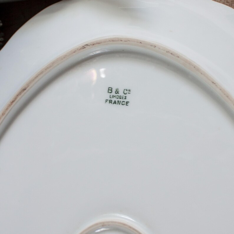 May include: White porcelain serving dish with a scalloped edge. The underside features the text "B & C&deg; LIMOGES FRANCE" in black lettering. The dish has a smooth, glossy surface and a slightly off-white hue.