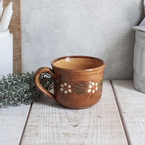May include: A brown ceramic mug with a handle, decorated with white flower and green leaf designs. The mug has a wide opening and a slightly textured surface. The mug is sitting on a white wooden surface.