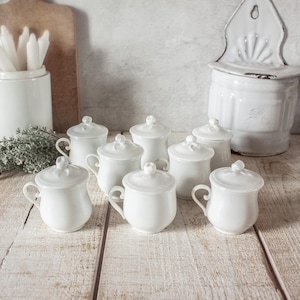 Könnte beinhalten: Eine Ansammlung weißer Keramikbecher mit Deckeln und Henkeln, angeordnet auf einer rustikalen Holzoberfläche. Die Becher haben ein klassisches Design und sind mit anderen weißen Küchenutensilien gruppiert.