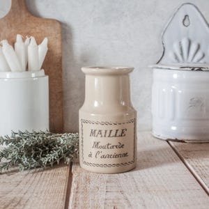 Puede incluir: Un tarro de cerámica beige con el texto "MAILLE Moutarde à l'Ancienne" en escritura negra. El tarro es cilíndrico con una base ligeramente más ancha y un cuello estrecho. Una ramita de hierbas verdes y otros recipientes blancos están en el fondo.