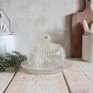 May include: A clear glass cloche with a decorative diamond pattern. The cloche is sitting on a clear glass plate. The cloche is on a white wooden surface.