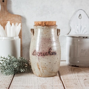 Può includere: Un vaso di ceramica rustico e maculato con coperchio in sughero, etichettato "Cornichons" in corsivo. Il vaso ha piccole maniglie e una combinazione di colori marrone chiaro e beige. Lo sfondo include un tagliere di legno e un contenitore bianco.