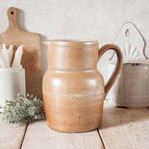 Peut inclure: Une cruche en grès beige avec une anse et un couvercle. La cruche a une surface texturée et est décorée d'une ligne pointillée sur le dessus. Une planche à découper en bois et des récipients en céramique blancs sont en arrière-plan.