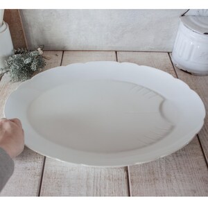 May include: A large, white, oval serving platter with a scalloped edge. The platter has a subtle leaf design on the surface. The background includes a wooden surface and a white container.