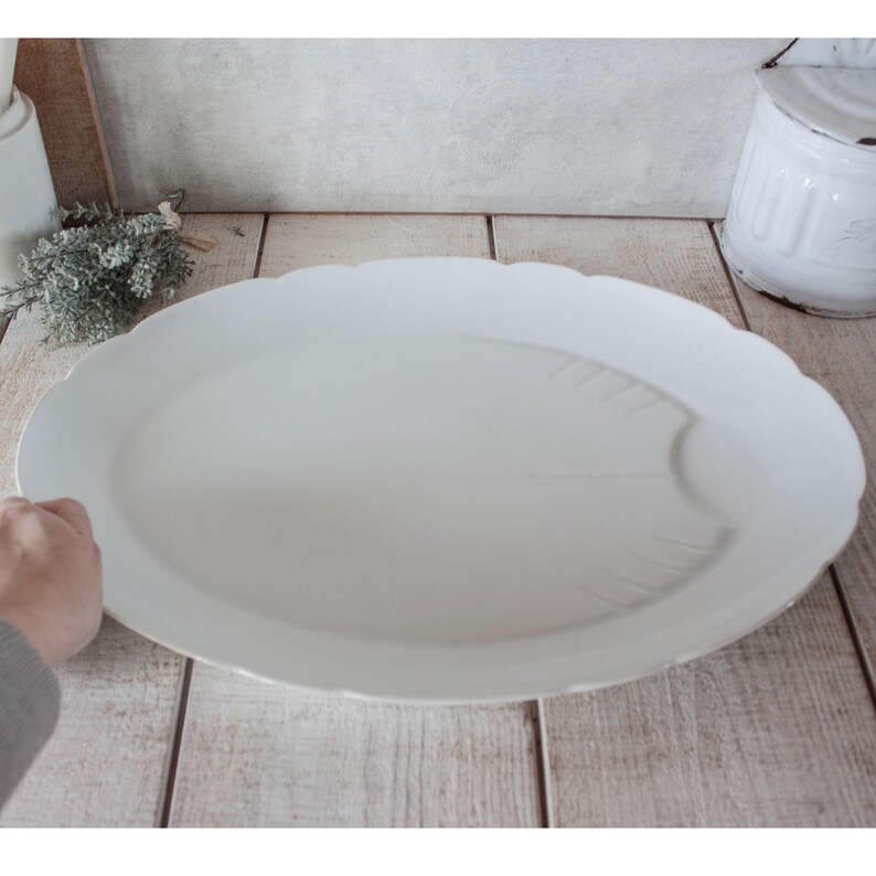 May include: A large, white, oval serving platter with a scalloped edge. The platter has a subtle leaf design on the surface. The background includes a wooden surface and a white container.