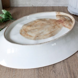 May include: A white, oval-shaped ceramic serving platter, viewed from the underside. The rim has a subtle, aged appearance. The center features a natural, textured pattern in shades of brown and tan. The platter is set against a wooden surface.