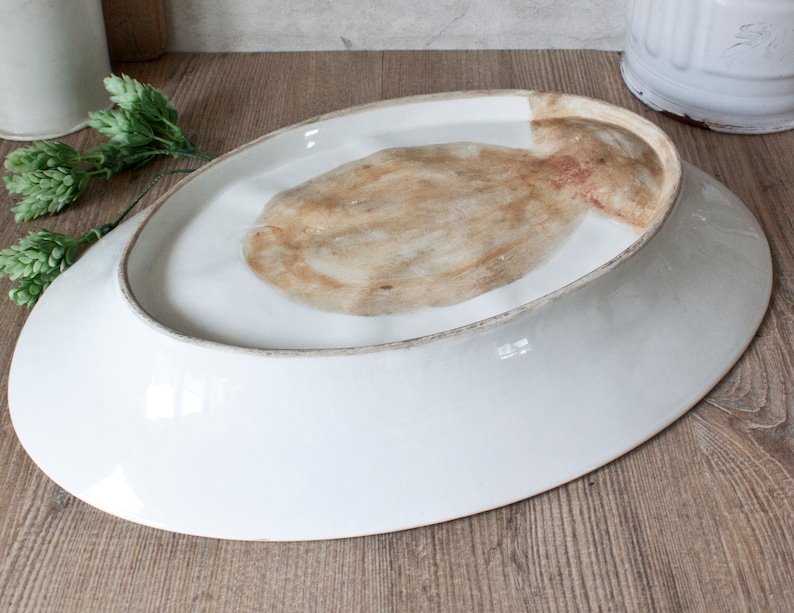 May include: A white, oval-shaped ceramic serving platter, viewed from the underside. The rim has a subtle, aged appearance. The center features a natural, textured pattern in shades of brown and tan. The platter is set against a wooden surface.