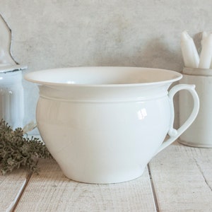 May include: White porcelain chamber pot with a rounded body and a single handle. The pot is sitting on a white wooden surface.