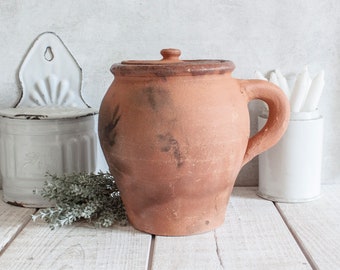 May include: A large, terracotta-colored pottery jar with a lid and a handle. The jar has a rustic, aged appearance with dark brown stains and is sitting on a white wooden surface.