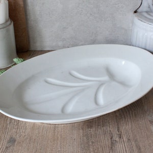 May include: A white oval serving platter with a raised leaf design. The platter is made of ceramic and sits on a wooden surface. The platter is approximately 16 inches long.