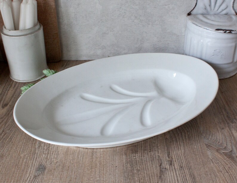May include: A white oval serving platter with a raised leaf design. The platter is made of ceramic and sits on a wooden surface. The platter is approximately 16 inches long.