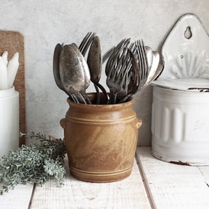 Peut inclure: Un assortiment de couverts anciens, comprenant des fourchettes et des cuillères, est présenté dans un pot en céramique marron. Le pot a deux petites poignées et est posé sur une surface en bois blanc. Un récipient blanc et un objet mural sont également visibles.