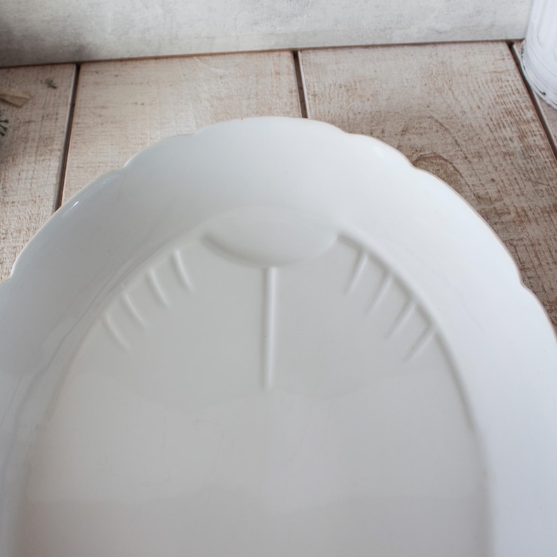 May include: A white, scalloped-edge serving dish. The dish has a central, raised design with vertical lines radiating outwards. The overall shape is oval, and the surface is smooth and glossy. The dish is made of ceramic or porcelain.