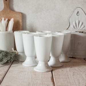 Könnte beinhalten: Sechs weiße Kelchgläser aus Keramik stehen auf einer hellen Holzoberfläche. Die Gläser haben eine Tulpenform und einen Sockel. Im Hintergrund befinden sich weitere weiße Keramikartikel, darunter ein Behälter mit Kerzen und ein Wandbehälter.