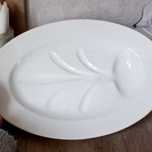 May include: A large, oval, white serving platter with a raised, leaf-like design in the center. The platter is held by a person's hand. A white ceramic container with white candles is visible in the background.