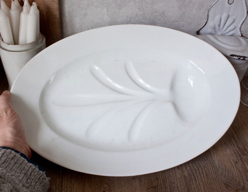 May include: A large, oval, white serving platter with a raised, leaf-like design in the center. The platter is held by a person's hand. A white ceramic container with white candles is visible in the background.