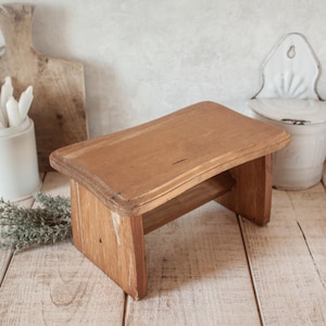 Puede incluir: Un pequeño taburete de madera rectangular con un acabado natural. El taburete tiene un diseño sencillo con cuatro patas y un estante inferior. La veta de la madera es visible y el taburete parece envejecido. Es una pieza de estilo rústico.