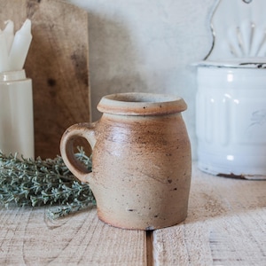 May include: A small, cylindrical stoneware mug with a thick, rounded rim and a single side handle. The exterior features a mottled, earthy tan and brown glaze with visible speckling and a textured, matte finish throughout the surface.
